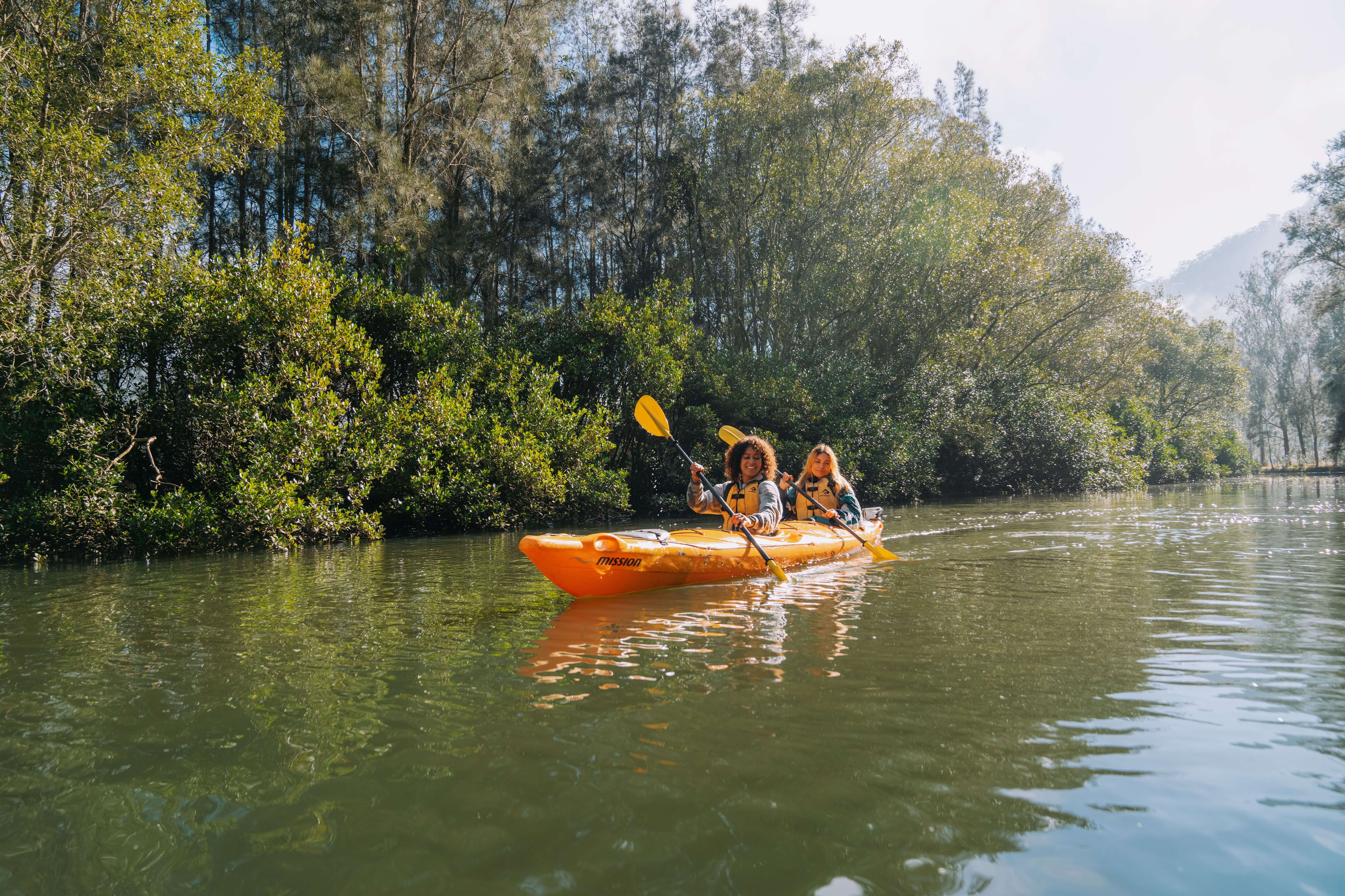 Picturesque view of an orange kayak with two people paddling down a river amongst green bushland. 