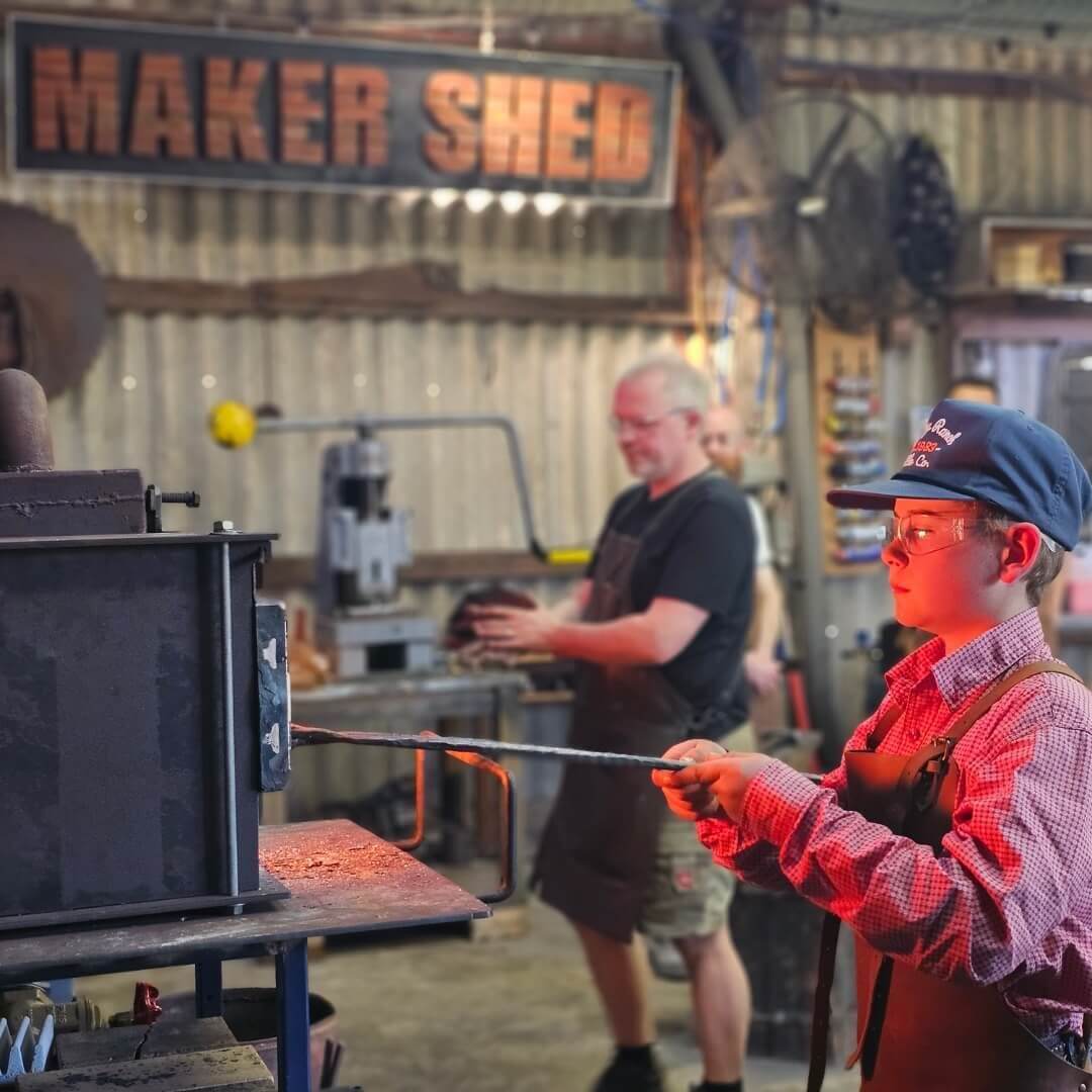 Standing in a tin maker's shed, a young boy stands wearing a red and white checkered shirt, holding a pair of tongs into a forge to heat a piece of metal, as a vibrant red glow from the fire is cast upon his face. Three other gentlemen stand in the background. 