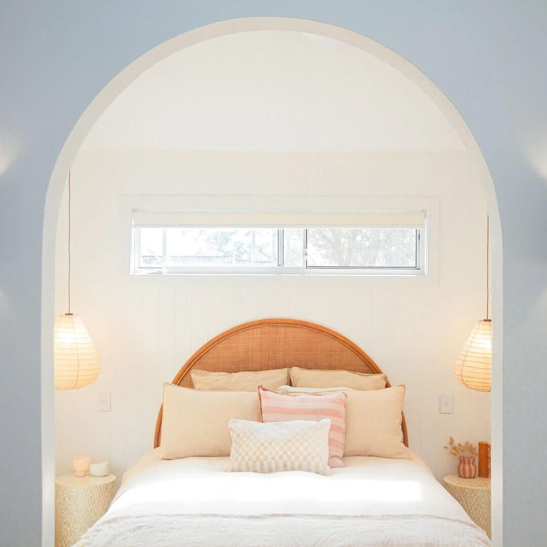 A stylish bedroom featuring a queen bed, warm toned bedding with six plush cushions, two circular bedside tables and a pendant light overhanging each bedside table.