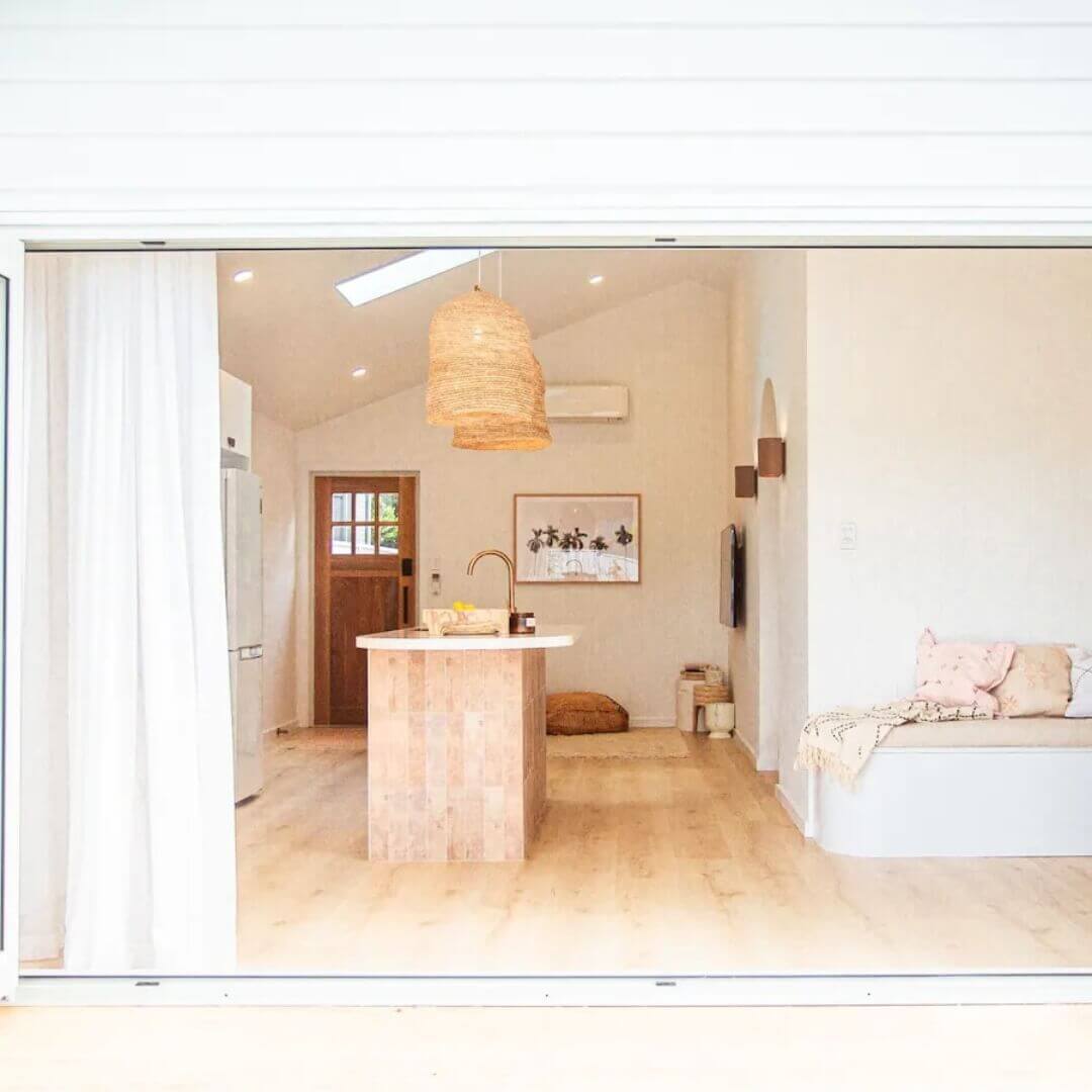 A sunlit open plan living space featuring two hand-woven pendant lights overhanging a stone kitchen island with a palm tree artwork and large dark wooden front door in the background, light timber floorboards and a white bench seat to the right of the island bench, adorned with a cream throw blanket and a couple of plush beige cushions. 