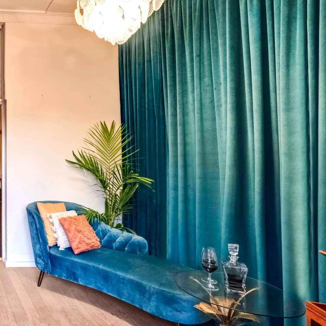 A cobalt blue chaise lounge adorned with cushions, a large vintage-style pendant and forest green crushed velvet curtains draping to the right of the lounge.