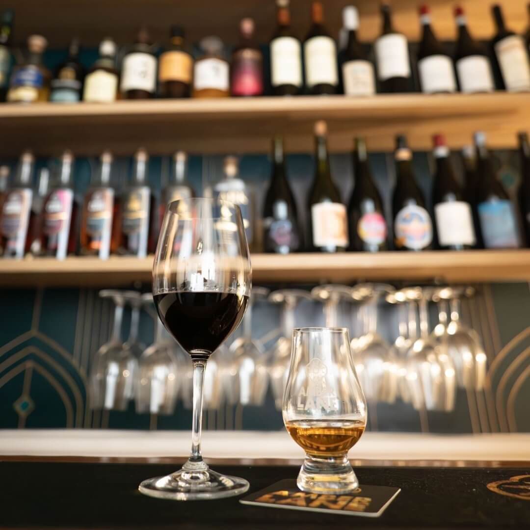 A glass of red wine and a neat glass of amber coloured whisky sitting on a bar with shelves of wine and whisky in the background.