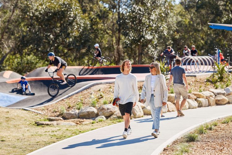 Three adults walking on a shared pathway, with a child on a BMX bike in a skate park in the background