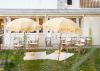 Outdoor picnic with yellow and white beach umbrellas with deck chairs, rugs and pillows on green grass in front of a white coastal styled restaurant.